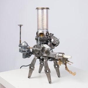 May include: A unique steampunk-style lamp constructed from repurposed metal components. The lamp features a cylindrical perforated shade, gears, and other mechanical parts, with a base of four legs. The lamp is silver and gray.