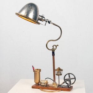 May include: A steampunk-style desk lamp with a silver dome shade, brass accents, and a wooden base. The lamp has a unique design with a small gear and a pencil holder.