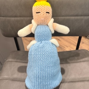 Puede incluir: Muñeca tejida a mano con forma de princesa. La muñeca tiene un vestido azul claro, una cara color melocotón y cabello amarillo. Los brazos y los detalles de la muñeca son blancos. La muñeca está sentada en una silla gris.