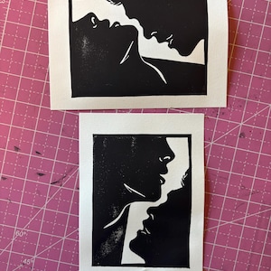 May include: Two black and white linocut prints. The top print shows two profiles in a kiss. The bottom print depicts three profiles in a close embrace. Both prints are on white paper, set against a pink cutting mat.