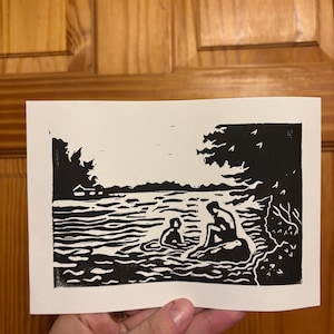 May include: Black and white linocut print depicting a serene lakeside scene. Two figures are in the water, with one seated on a rock. Trees and foliage frame the water's edge, creating a tranquil landscape.
