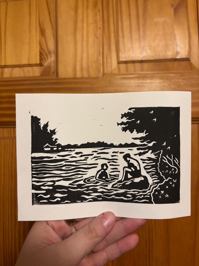 May include: Black and white linocut print depicting a serene lakeside scene. Two figures are in the water, with one seated on a rock. Trees and foliage frame the water's edge, creating a tranquil landscape.