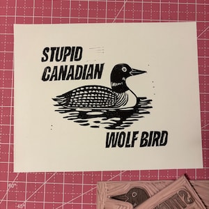 May include: A black and white print on white paper featuring a stylized illustration of a loon on water. The text reads "STUPID CANADIAN" above the bird and "WOLF BIRD" below.