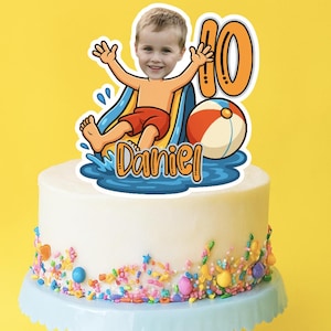May include: A white frosted cake with colorful sprinkles on a light blue cake stand. A cake topper features a boy on a waterslide, the number 10, and the name "Daniel". A beach ball is also included in the topper.