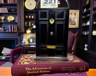 以下が含まれることがあります： 金色のノッカーが付いた黒いミニチュアのドアと「221B」の住所が3冊の本の上に置かれています。「シャーロック・ホームズの冒険」、「シャーロック・ホームズの帰還」、そして「バスカヴィル家の犬」というタイトルの本です。