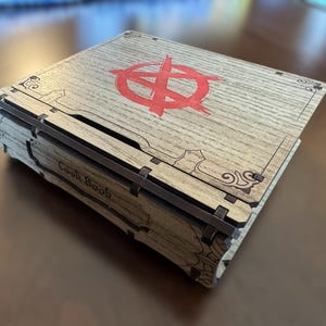 Puede incluir: Caja de libro de cocina de madera con un símbolo rojo en la tapa. La caja tiene un acabado de veta de madera clara y las palabras "Cook Book" son visibles en el lateral. La caja está cerrada y se encuentra sobre una superficie marrón.