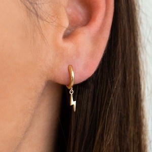 Puede incluir: Pendientes de aro de oro con un pequeño dije de rayo colgando de cada aro.