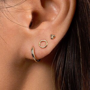 May include: A close-up of a person's ear with three gold earrings. The earrings are a small circle, a larger circle, and a cluster of three small gold balls.