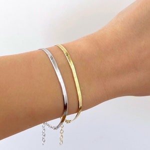 May include: Two thin bracelets on a wrist. One is a gold snake chain bracelet and the other is a silver snake chain bracelet with a small silver chain dangling from it.