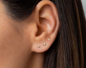 May include: A close-up of a person's ear with three small diamond stud earrings. The earrings are set in gold and are worn in the lobe and helix piercings.