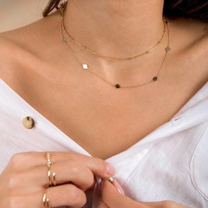 Puede incluir: Una mujer con una camisa blanca y un collar de cadena dorada con peque&ntilde;os abalorios redondos y cuadrados. Lleva varios anillos de oro en los dedos.
