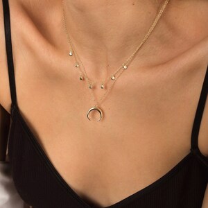 Puede incluir: Un collar de oro con un colgante de luna creciente y una cadena delicada con peque&ntilde;os colgantes en forma de l&aacute;grima.