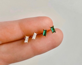 May include: Three pairs of gold stud earrings with rectangular gemstones. One pair features a clear gemstone, while the other two pairs feature green gemstones.