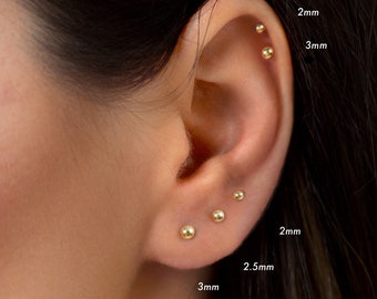 Puede incluir: Primer plano de la oreja de una persona con varios pendientes de bola de oro de diferentes tamaños. Los pendientes están etiquetados con sus tamaños en milímetros: 2 mm, 2,5 mm, 3 mm.