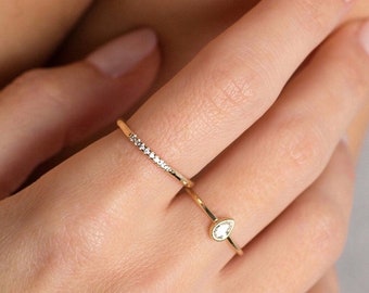 May include: Two gold rings on a finger. The top ring features a single pear-shaped diamond. The bottom ring has a row of small diamonds.