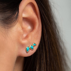 May include: Gold earrings with three turquoise teardrop-shaped stones. The earrings are arranged in a curved line.