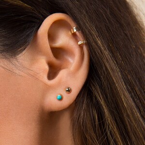 May include: A close-up of a person's ear with three gold earrings. The earrings are a small turquoise stone, a small gold ball, and a gold hoop with small diamonds.