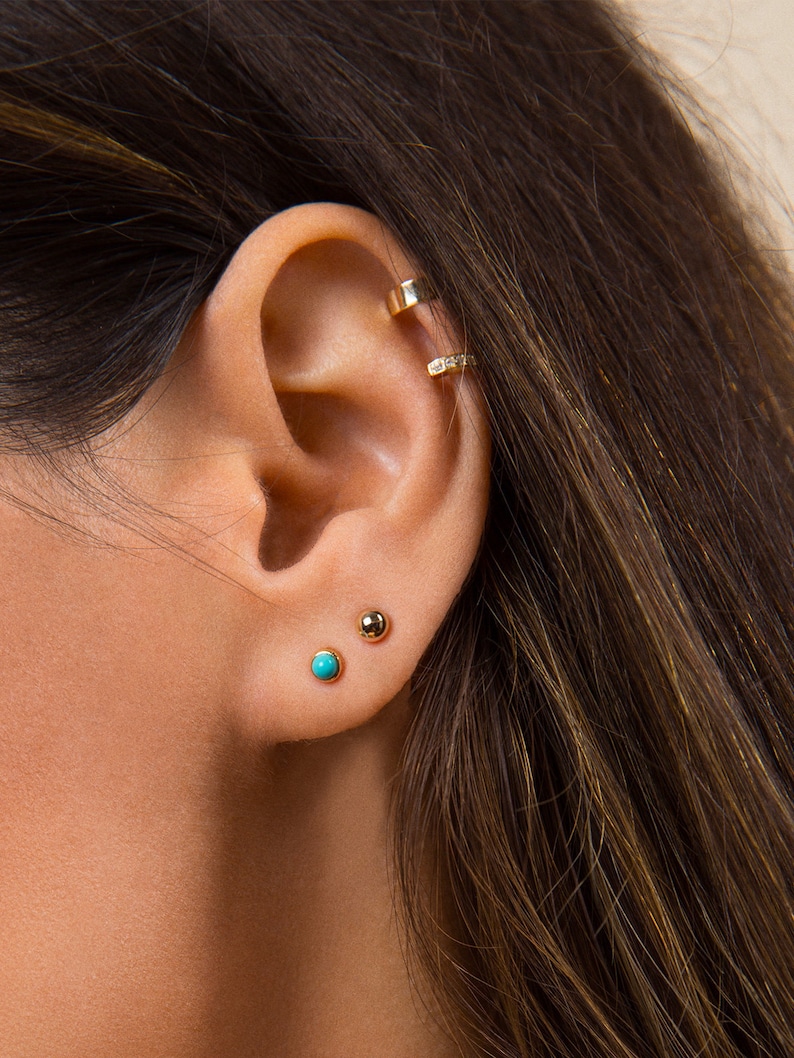 May include: A close-up of a person's ear with three gold earrings. The earrings are a small turquoise stone, a small gold ball, and a gold hoop with small diamonds.