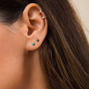 May include: A close-up of a person's ear with three small gold earrings. The earrings are a 4mm hoop, a small gold ball, and a turquoise gemstone.