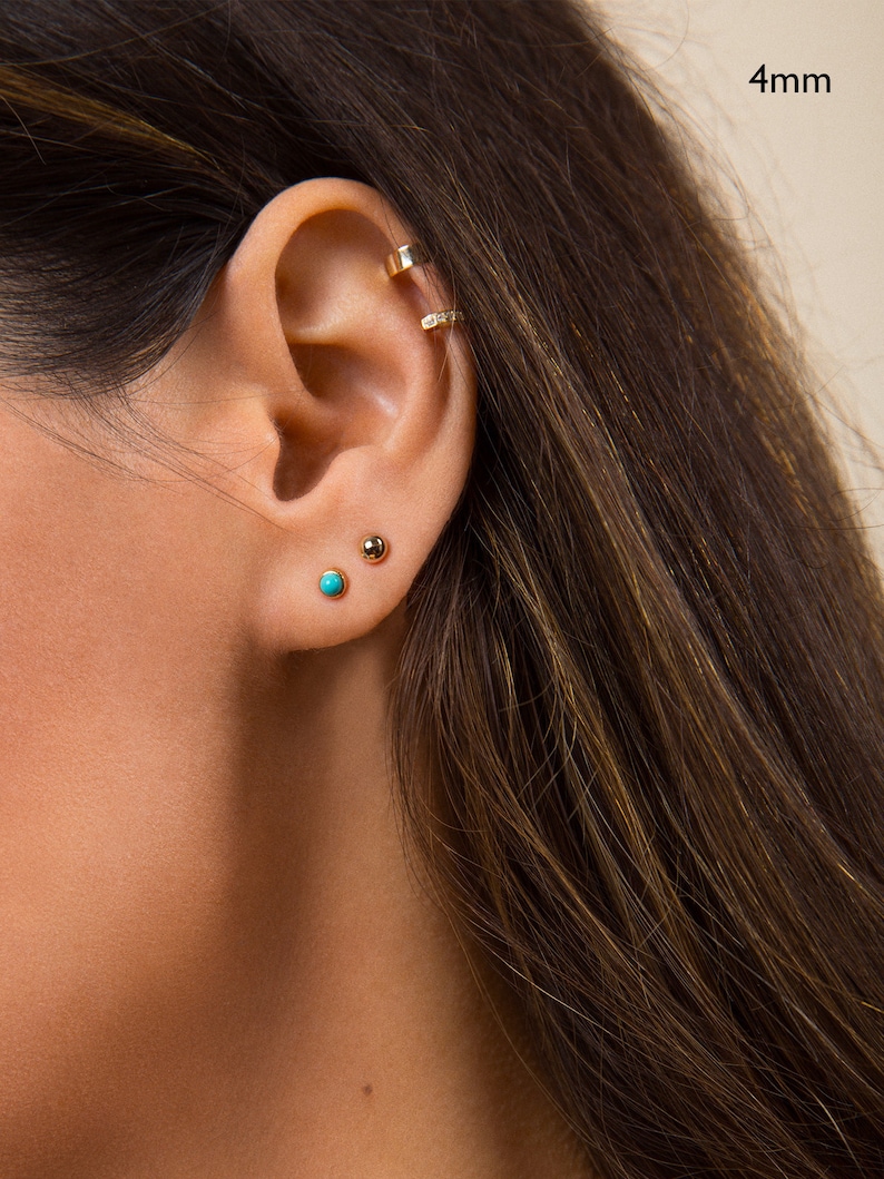 May include: A close-up of a person's ear with three small gold earrings. The earrings are a 4mm hoop, a small gold ball, and a turquoise gemstone.