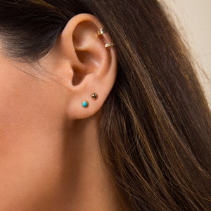 May include: Close-up of a person's ear with two small earrings. The earring on the lower lobe is turquoise blue, and the earring on the upper lobe is gold. The text "4mm" is visible in the upper right corner of the image.