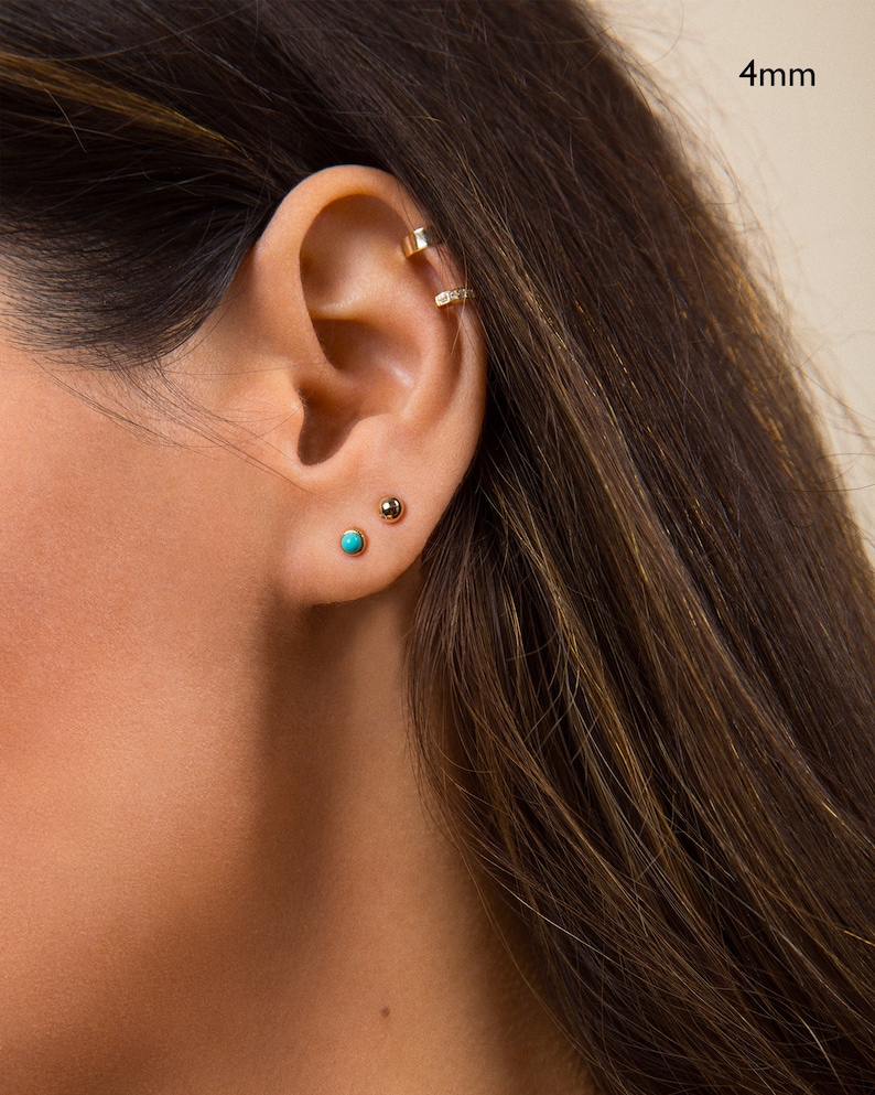 May include: Close-up of a person's ear with two small earrings. The earring on the lower lobe is turquoise blue, and the earring on the upper lobe is gold. The text "4mm" is visible in the upper right corner of the image.