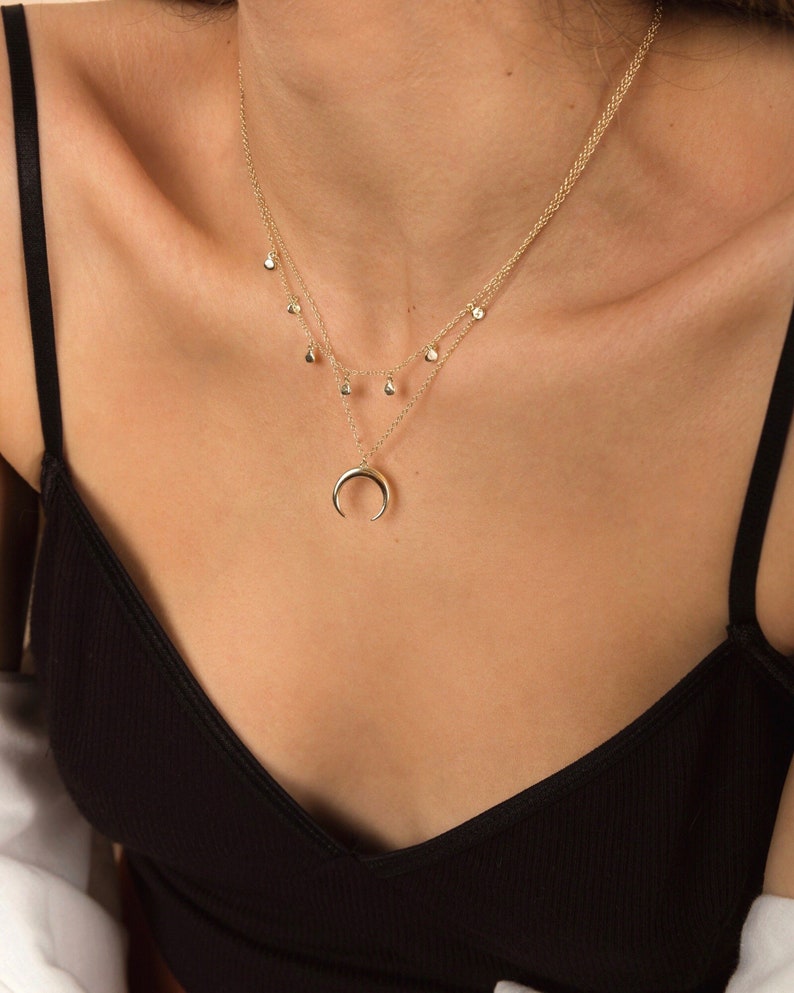 Puede incluir: Un collar de oro con un colgante de luna creciente y una cadena delicada con peque&ntilde;os colgantes en forma de l&aacute;grima.