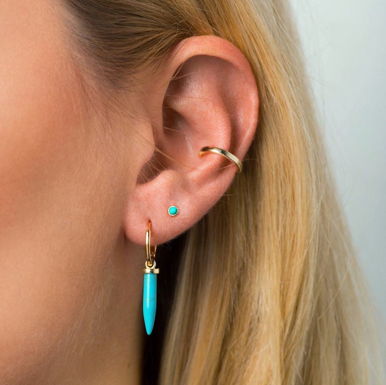 May include: Gold hoop earrings with a turquoise stone dangle. The earrings are worn in the lower lobe of the ear. The ear also has a gold ear cuff and a small turquoise stud.