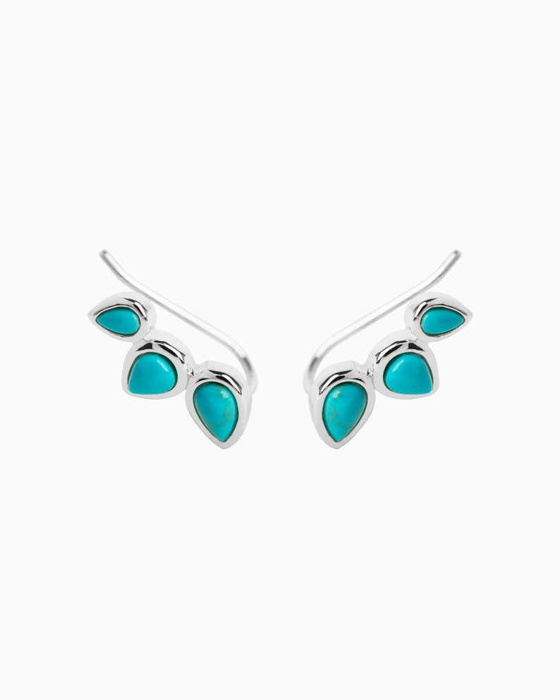 May include: A pair of silver ear cuffs with turquoise stones. The cuffs are curved and have three stones each.