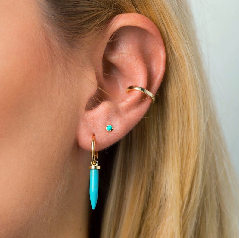 May include: A close-up of a person's ear with three gold earrings. The bottom earring is a turquoise teardrop-shaped dangle earring. The middle earring is a small turquoise stud. The top earring is a gold hoop.