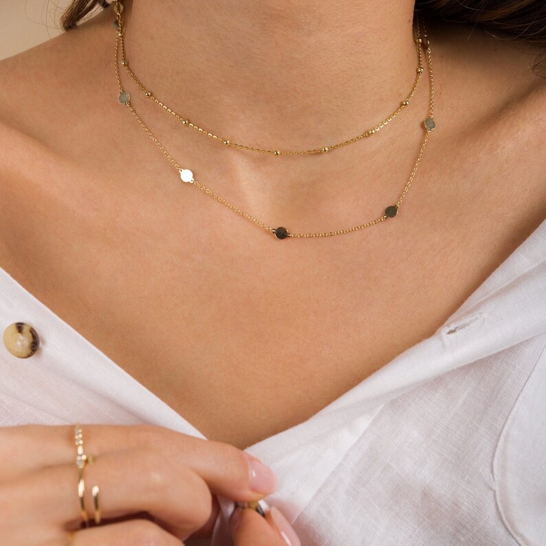 Puede incluir: Un collar de cadena de oro con peque&ntilde;os dijes redondos. El collar est&aacute; superpuesto con una segunda cadena m&aacute;s fina con peque&ntilde;as cuentas.