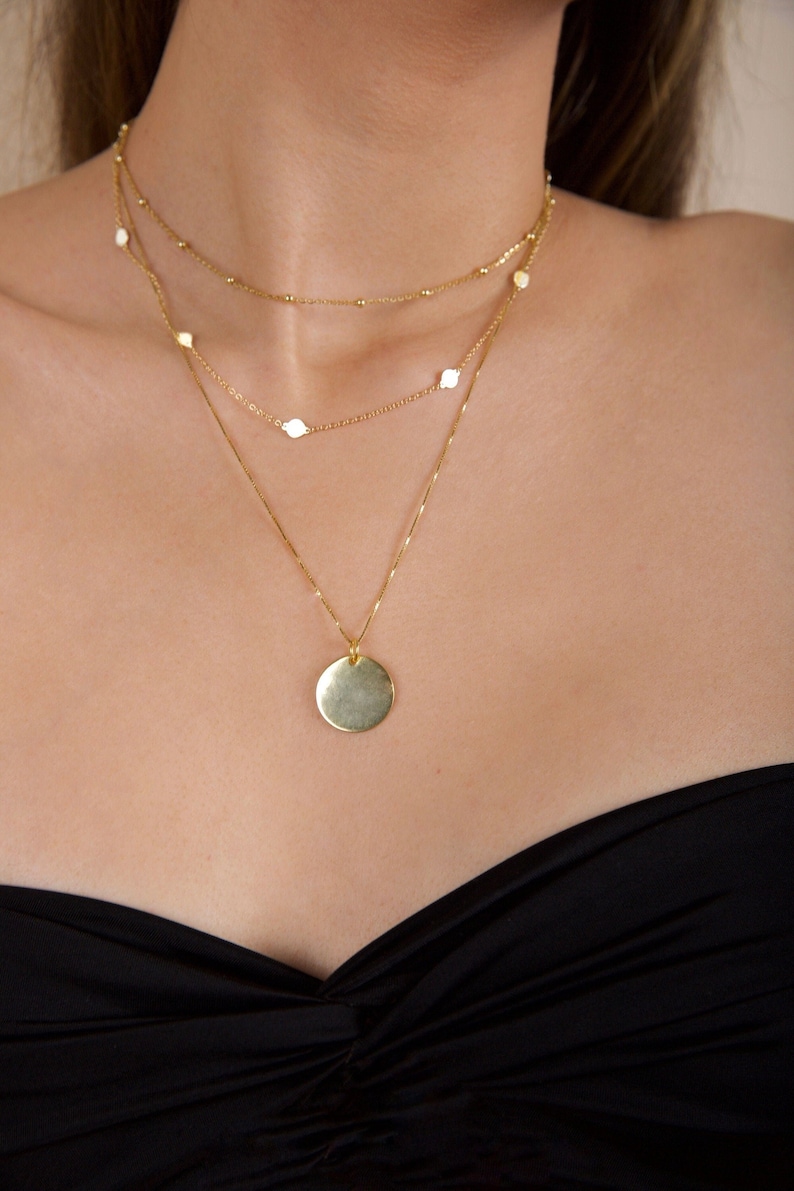 Puede incluir: Un collar de oro con un colgante redondo y una cadena m&aacute;s peque&ntilde;a con peque&ntilde;os abalorios redondos. El collar lo lleva una persona con un top negro sin tirantes.