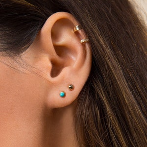 May include: A close-up of a person's ear with three gold earrings. The earrings are small and round, with one featuring a turquoise stone.