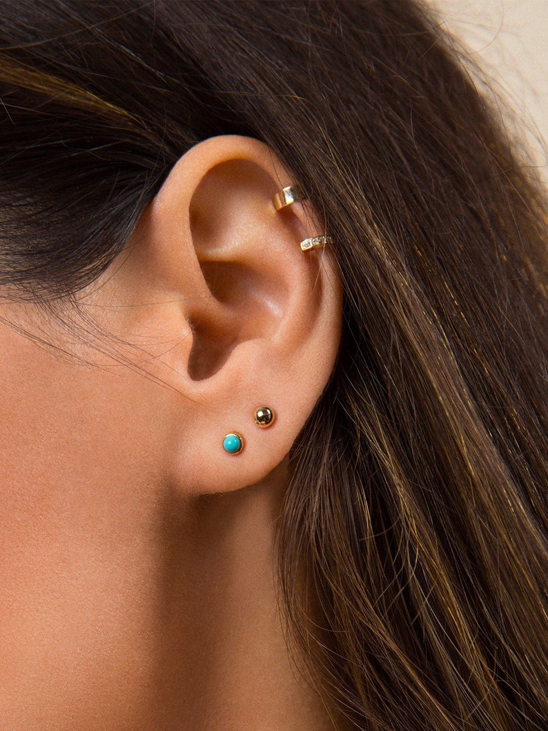 May include: A close-up of a person's ear with three gold earrings. The earrings are small and round, with one featuring a turquoise stone.