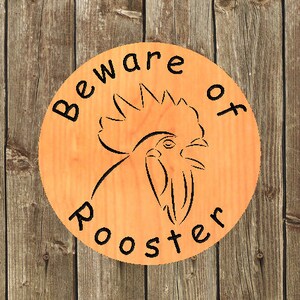 Puede incluir: Un letrero redondo de madera con el texto "Beware of Rooster" y una silueta negra de la cabeza de un gallo.