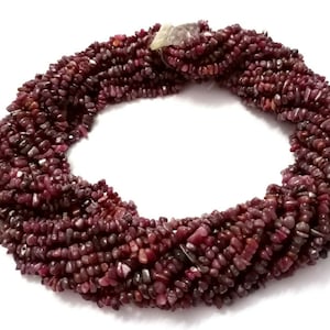 Peut inclure: Un collier composé de plusieurs brins de petites pierres précieuses rouge foncé de forme irrégulière. Les pierres ont un aspect légèrement poli et sont étroitement tassées, créant une surface texturée. Le collier est présenté sur fond blanc.