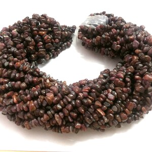 May include: A necklace composed of numerous small, irregularly shaped, dark red-brown gemstones. The stones are strung together, forming a thick, multi-strand necklace. The gemstones have a polished appearance and reflect light, creating a subtle shimmer.