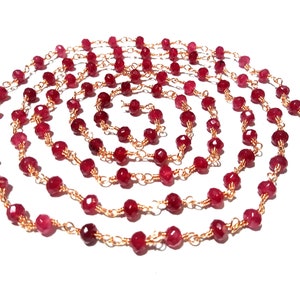 May include: A necklace composed of small, faceted, deep red gemstones strung on delicate gold-colored wire. The necklace is arranged in a spiral pattern against a white background, showcasing the jewelry's design.