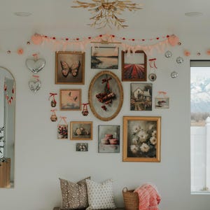 May include: A white wall gallery with a variety of framed artwork, including landscapes, still lifes, and a butterfly. The gallery is decorated with a pink and red garland and a gold chandelier. A wooden bench with two pillows and a basket sits in front of the gallery.