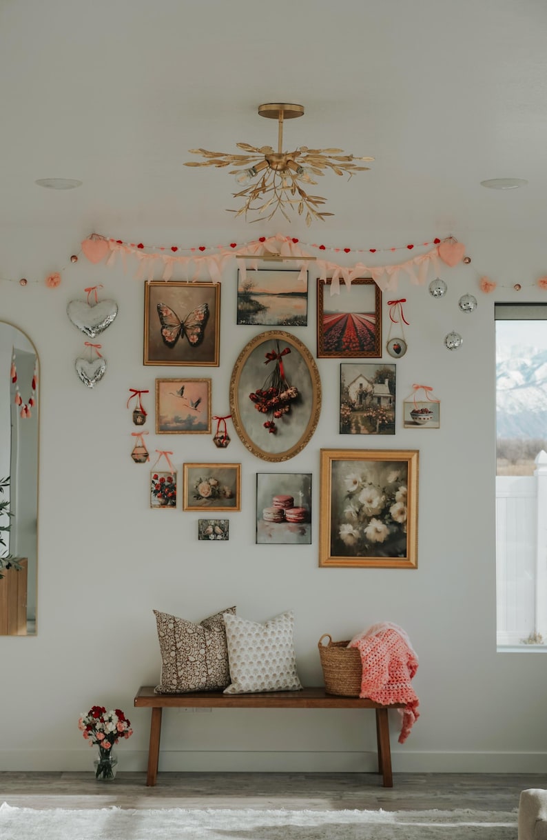 May include: A white wall gallery with a variety of framed artwork, including landscapes, still lifes, and a butterfly. The gallery is decorated with a pink and red garland and a gold chandelier. A wooden bench with two pillows and a basket sits in front of the gallery.