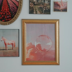 May include: A gallery wall with various framed artwork, including a pink horse, a house in a garden, a field of tulips, and a bubble floating in a pink sky.
