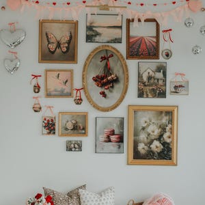 May include: A white wall gallery featuring a variety of framed artwork, including floral prints, landscapes, and a butterfly. The gallery is decorated with a pink and red heart garland and a wooden bench with two pillows and a basket.