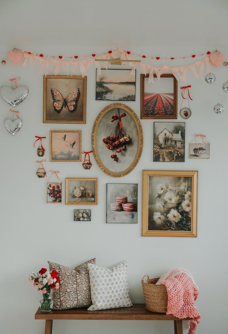 May include: A white wall gallery featuring a variety of framed artwork, including floral prints, landscapes, and a butterfly. The gallery is decorated with a pink and red heart garland and a wooden bench with two pillows and a basket.