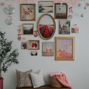 May include: A gallery wall with a variety of framed artwork, including landscapes, still lifes, and abstract pieces. The wall is decorated with a pink and red garland of hearts. A wooden bench with pillows and a blanket sits in front of the wall.