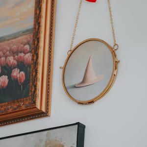 May include: A round, gold-framed picture with a white background and a pink witch's hat hanging from a gold chain with a red ribbon.