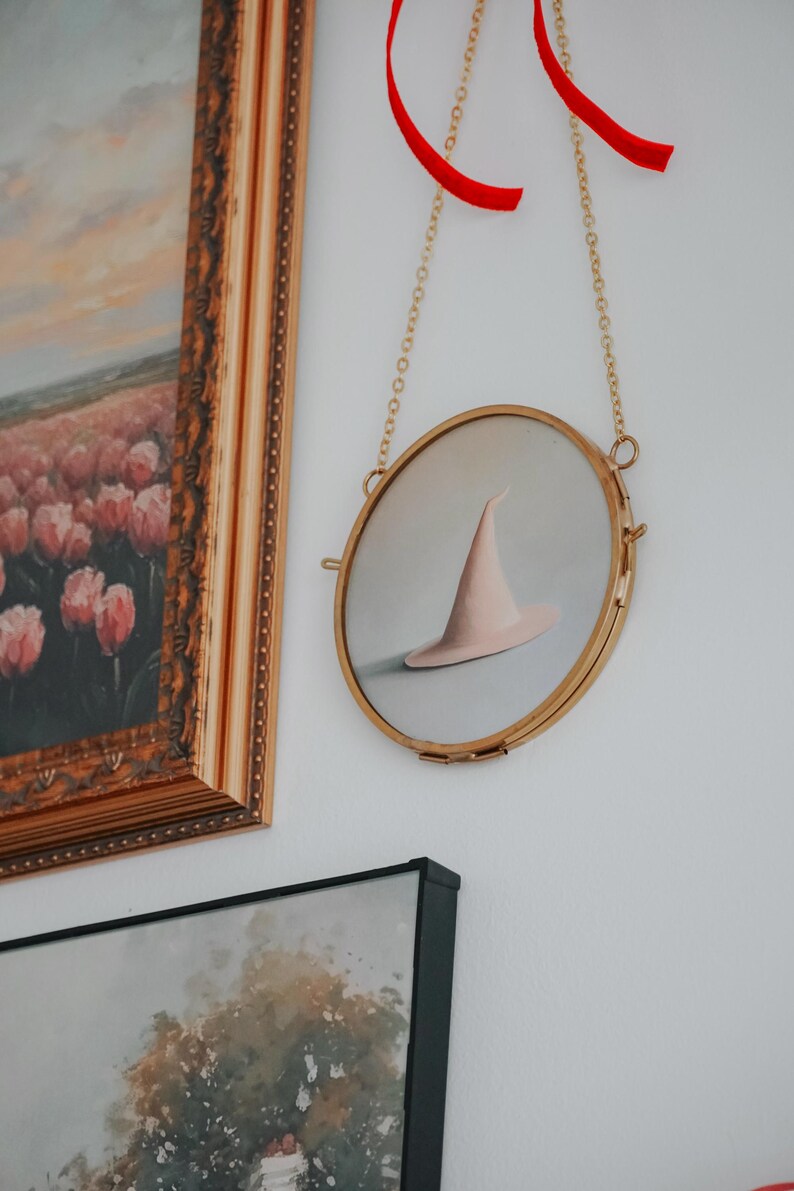 May include: A round, gold-framed picture with a white background and a pink witch's hat hanging from a gold chain with a red ribbon.