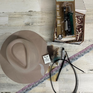 May include: A craft kit for making a hat with a brown felt hat, a black leather cord, a white feather, a black marker, a grey and pink patterned ribbon, and a woodburning tool in a box.