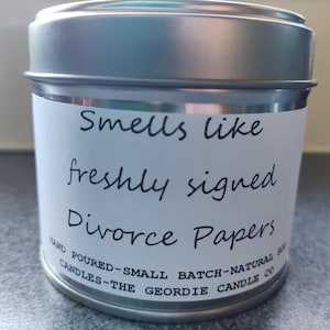May include: A silver tin candle with a white label that reads "Smells like freshly signed Divorce Papers". The label also includes the text "Hand Poured - Small Batch - Natural Soy Candles - The Geordie Candle Co."