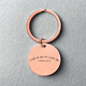 Op de afbeelding: Roségouden sleutelhanger met een ronde hanger. De hanger heeft de gegraveerde tekst "BE HOLY AS HE IS HOLY" en "1 PETER 1:14-15". De sleutelring is met een kleine lus aan de hanger bevestigd.