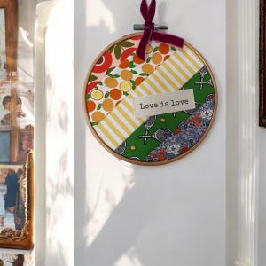 May include: A round embroidery hoop with a colorful patchwork design. The fabric is a mix of patterns, including oranges, lemons, stripes, and floral designs. The text "Love is love" is embroidered on a white fabric patch.
